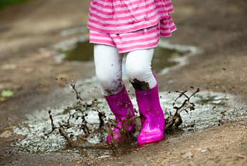 Una niña saltando con botas de goma de color rosa en un charco de barro.